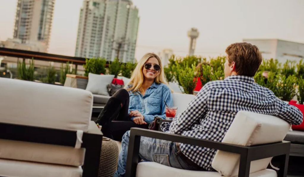 A couple enjoying drinks on the HG Sply Co rooftop lounge in Lower Greenville with the Dallas skyline in the background.
